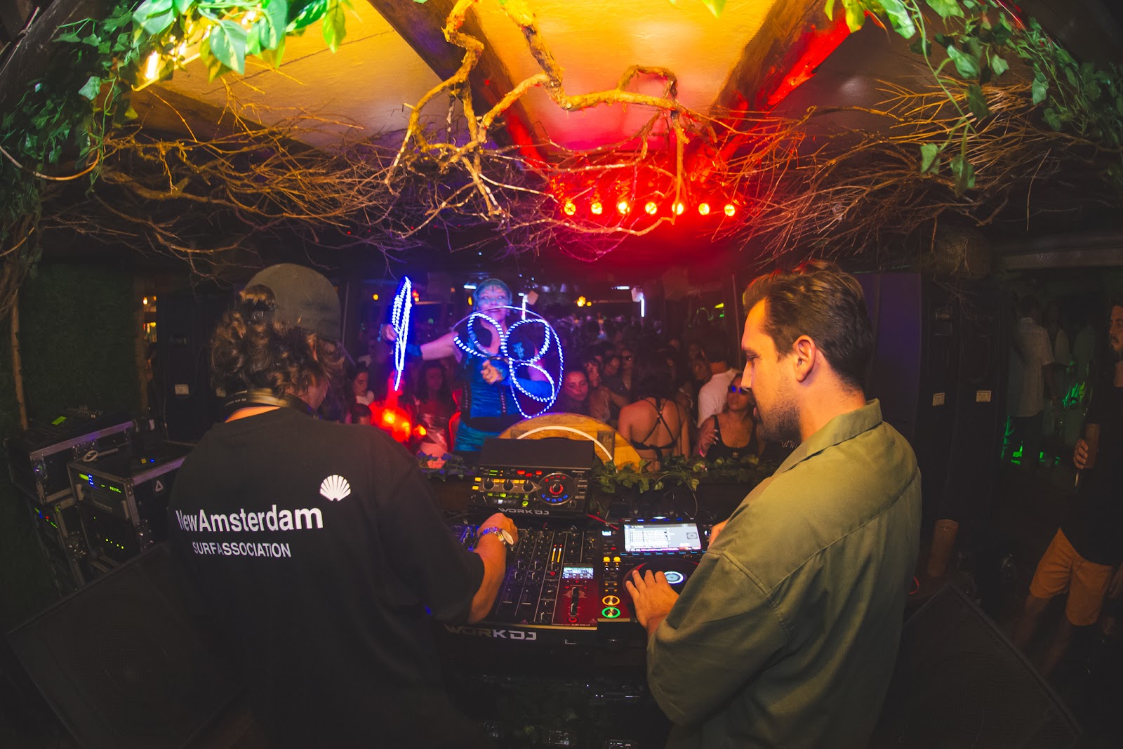 Os DJs na cabine e a pista dançando à frente, sob a madeira e o cipó